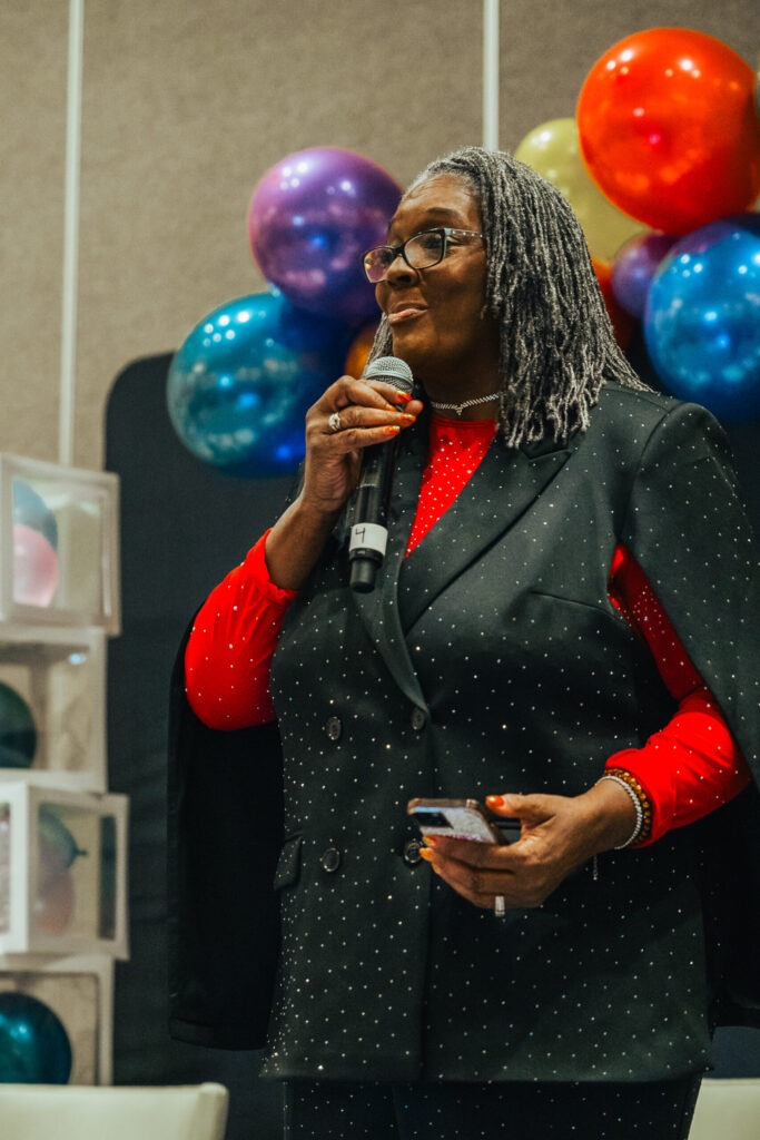 A speaker, dressed in a sparkling black blazer over a red top, holds a microphone and addresses an audience