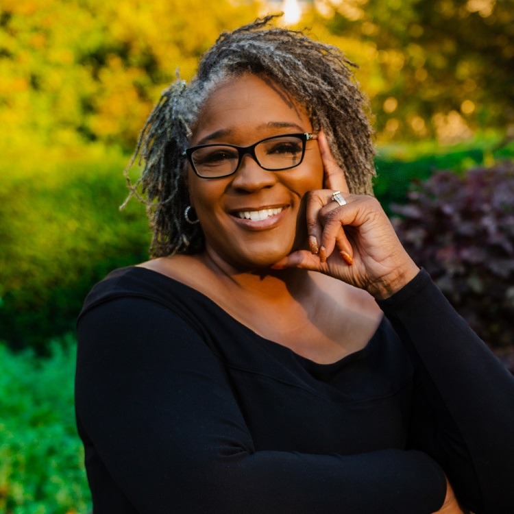 Smiling woman with glasses and gray hair poses thoughtfully in a lush green garden
